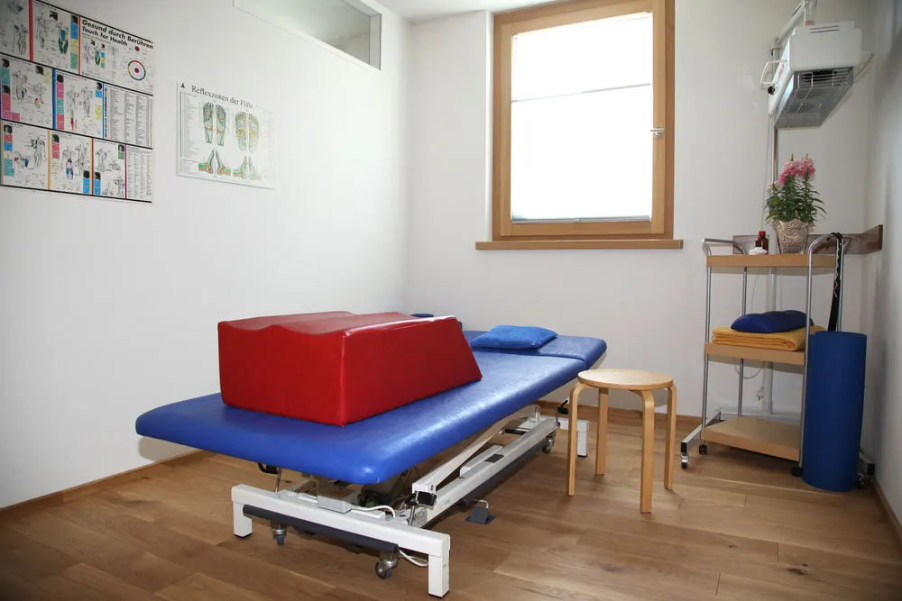 Behandlungsraum mit blauer Liege, Holzfenster und Hocker