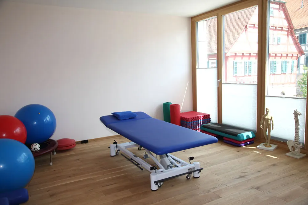 Heller Therapieraum mit Liege, Gymnastikbällen und großen Holzfenstern